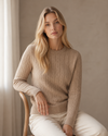 Woman wearing a beige cable-knit sweater sitting on a chair against a neutral wall.