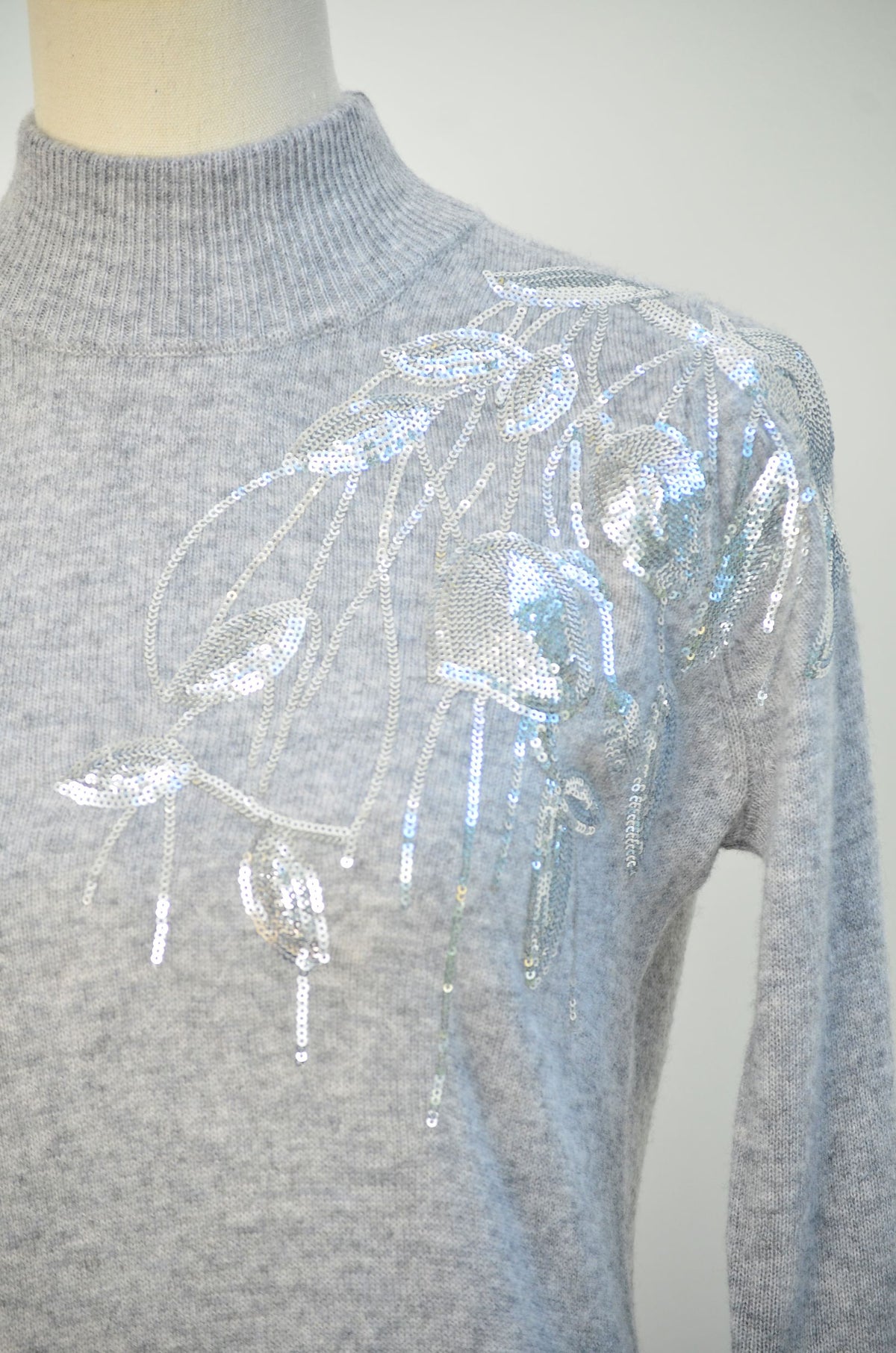 A close-up of a light sequin embellished dress displayed on a mannequin, featuring a large decorative sequin and bead embellishment in silver and pale blue across the upper chest and shoulder.