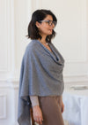 A woman with short dark hair and glasses wearing a gray cowl-neck cashmere wool shawl over a beige top and brown pants, standing in a bright white interior room.