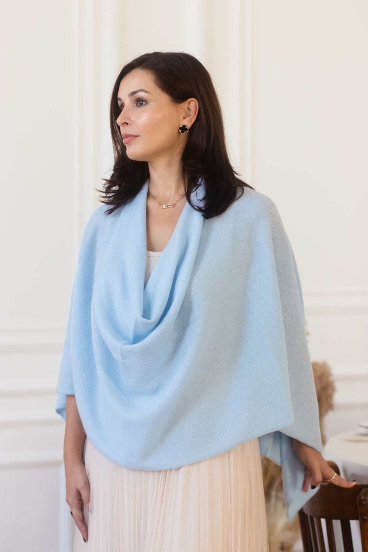 A woman wearing a light blue draped cashmere wool shawl over a cream-colored top and skirt, standing in a bright indoor setting with white wall molding.