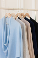 A row of lightweight draped knit shawls hanging on wooden hangers from a light wood clothing rack, shown in light blue, beige, taupe, navy, and black.
