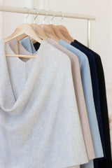 Four women's knit cowl-neck cashmere shawls in light gray, beige, pale blue, and navy blue, hanging on wooden hangers from a white clothing rack.