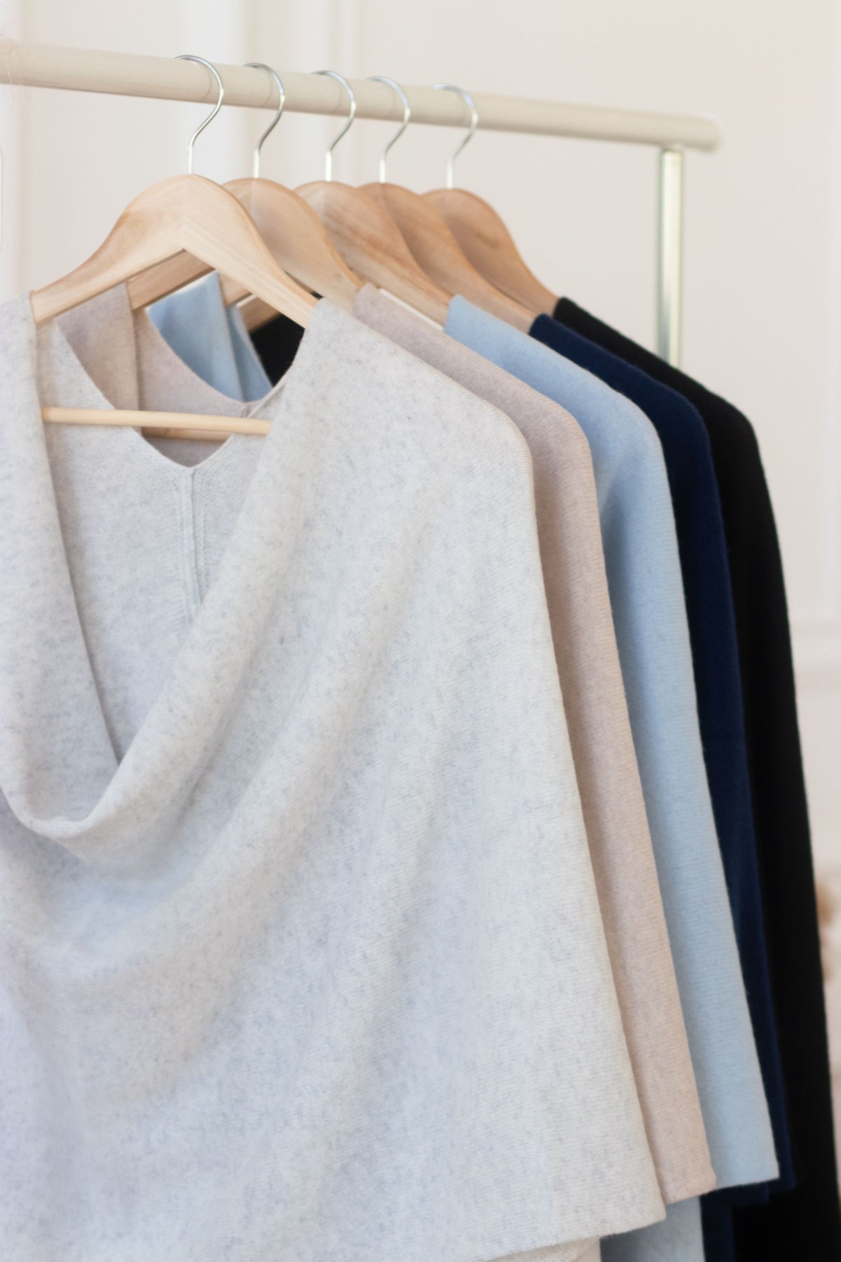 Four women's knit cowl-neck cashmere shawls in light gray, beige, pale blue, and navy blue, hanging on wooden hangers from a white clothing rack.