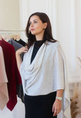 A woman wearing a light gray draped cowl giant cashmere scarf over a black top and black skirt, standing in a boutique next to a clothing rack.