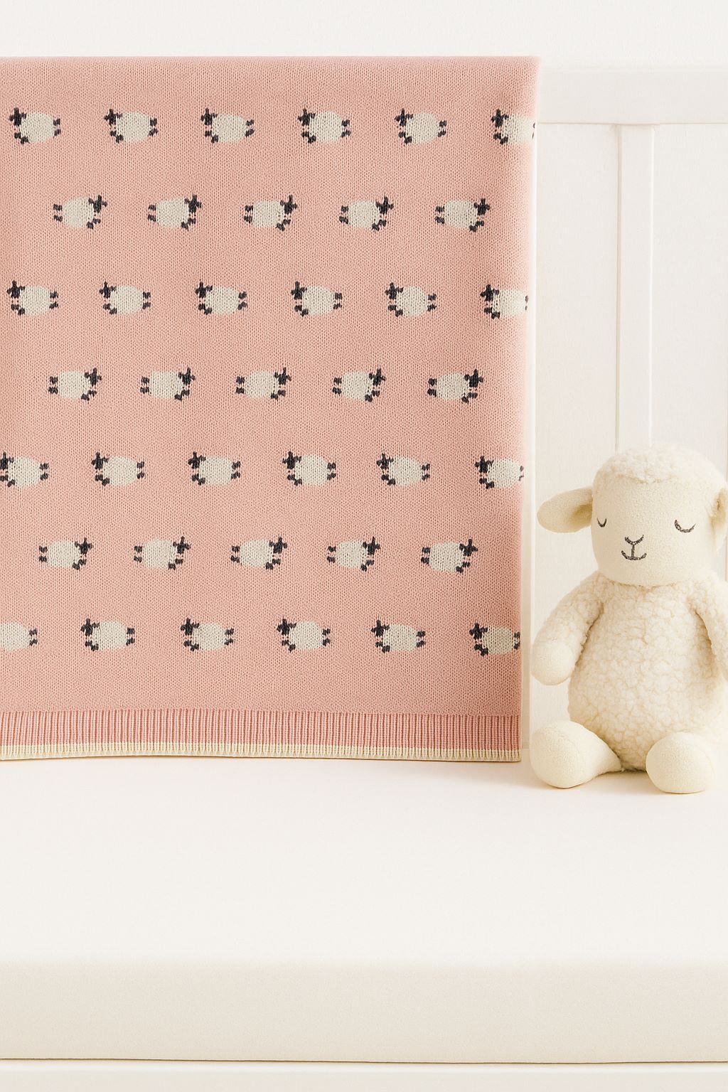 A pink knitted blanket with a repeating black-and-white sheep pattern, draped over a white crib next to a plush cream-colored stuffed sheep.