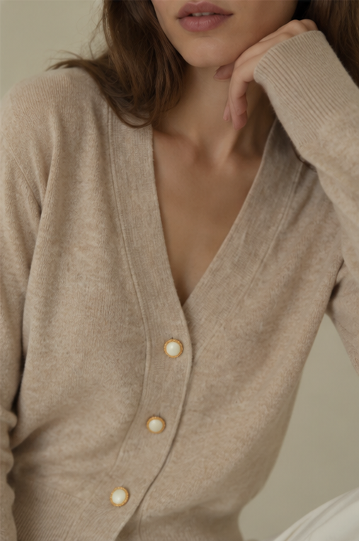 A close-up of a woman wearing a beige knit cardigan with large cream-colored buttons, styled in a soft, warm indoor setting.