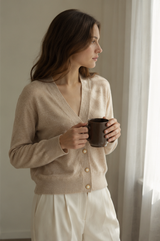 A woman in a beige knit cardigan and white pants holds a dark brown ceramic mug, standing in a softly lit room near a window with sheer curtains.