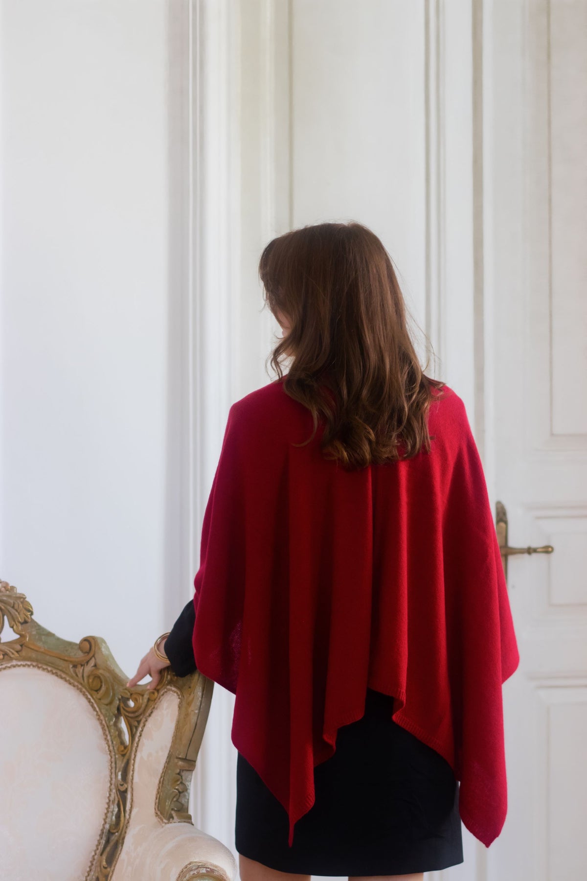 A woman viewed from behind wearing a bright red draped cashmere wool shawl over a black skirt, standing indoors near a white door and a cream-colored ornate chair.