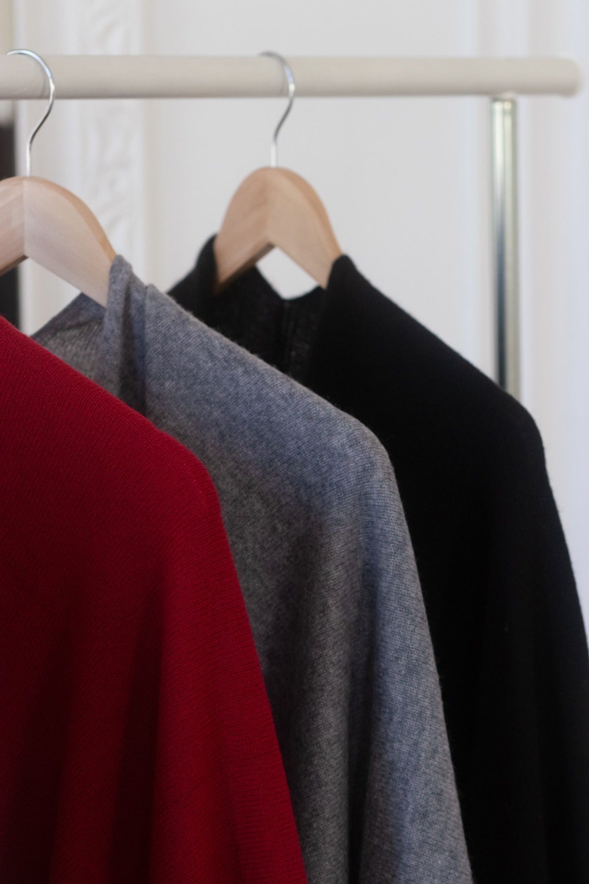 Three knit sweaters in red, gray, and black hanging on wooden hangers from a light wood clothing rack.