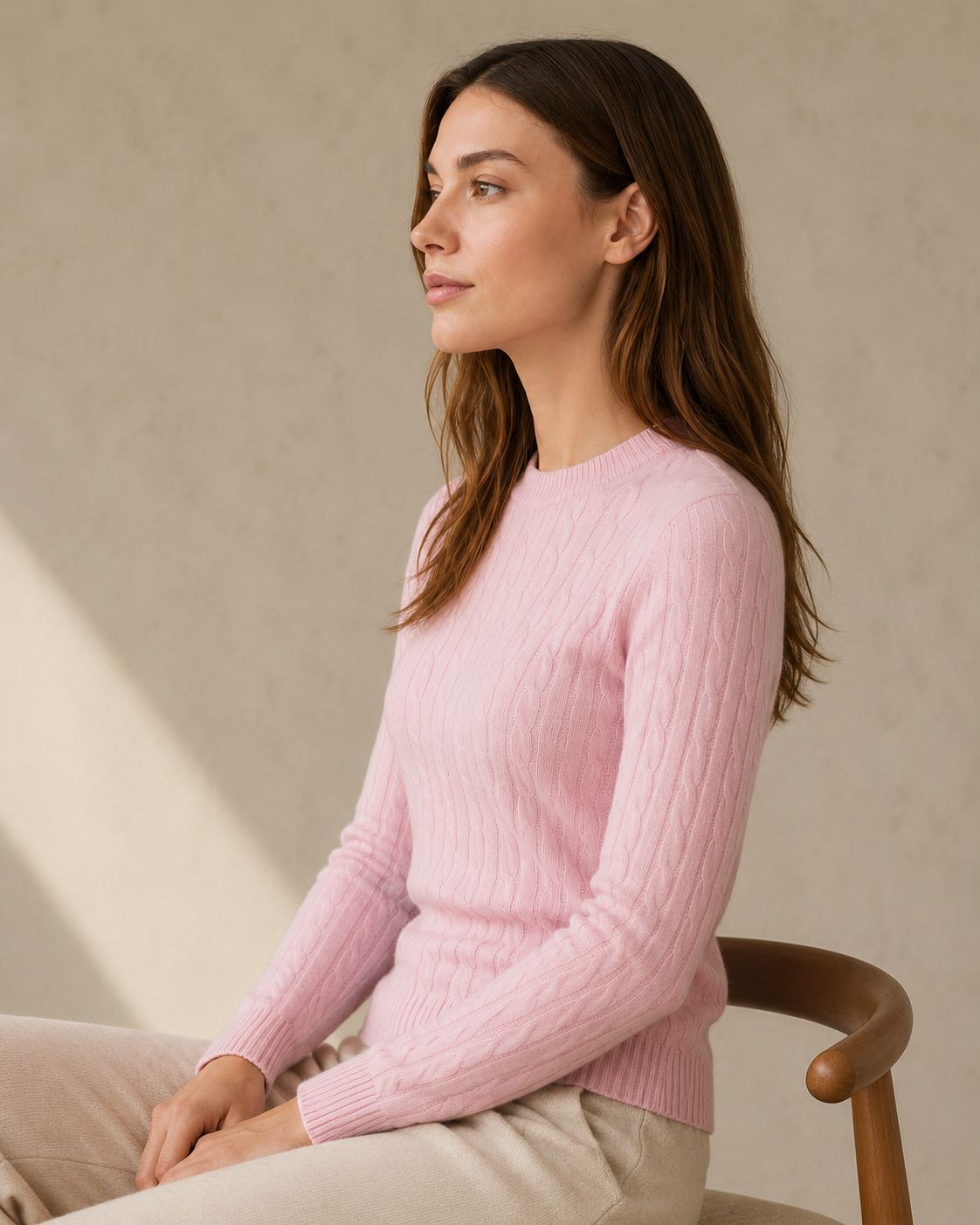 Woman wearing a pink cashmere wool cable crewneck sweater sitting on a chair against a beige wall