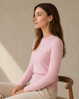 Woman wearing a pink cashmere wool cable crewneck sweater sitting on a chair against a beige wall
