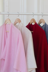 Four lightweight draped cashmere shawls or wraps in pink, cream, deep red, and blue-gray hanging on wooden hangers from a white clothing rack.