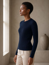 Woman wearing a navy blue cotton cable crewneck sweater and beige pants standing in a softly lit room.