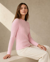 Woman wearing a pink cashmere wool cable crewneck sweater sitting on a light-colored surface with a neutral background