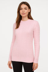 A woman wearing a light pink cable-knitcashmere crewneck sweater and black pants, standing against a plain white studio background.