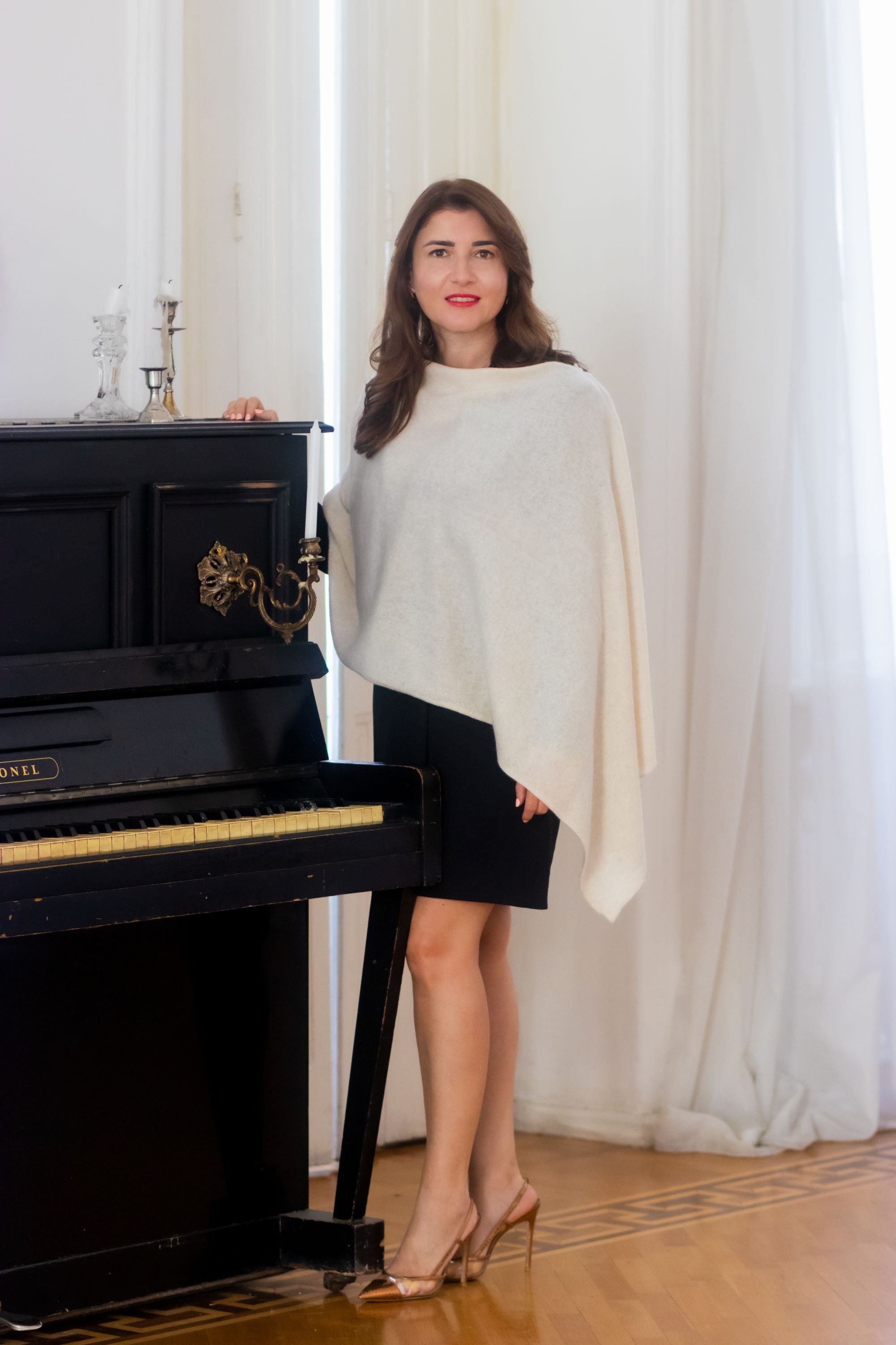 A woman stands in a bright room next to a black upright piano, wearing a cream-colored asymmetrical draped cashmere wool shawl, a black skirt, and beige high heels.