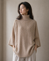 Woman wearing a beige cable turtleneck tunic knit sweater against a neutral background