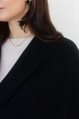 Close-up of a woman wearing a delicate gold bar necklace with a small engraved nameplate, styled with a black cashmere wool cardigan and a light gray top.