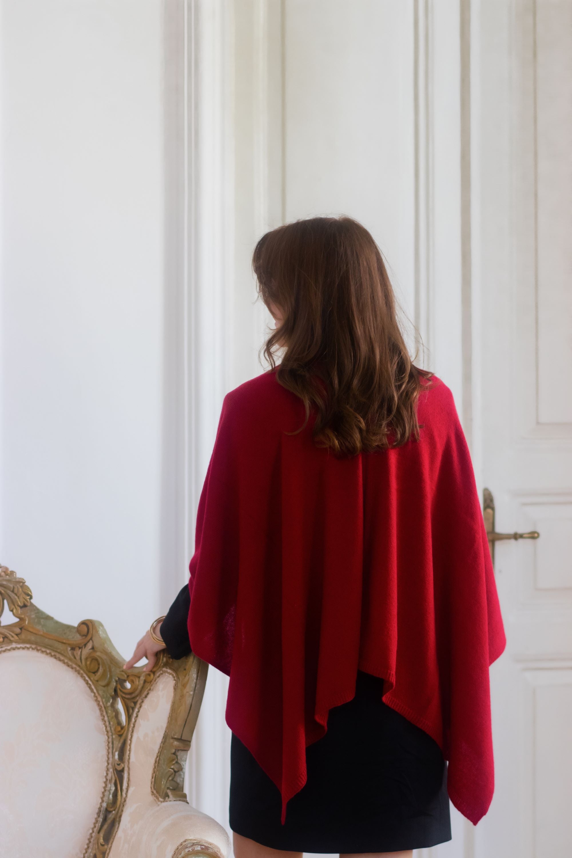 A woman viewed from behind wearing a bright red draped cashmere wool shawl over a black skirt, standing indoors near a white door and a cream-colored ornate chair.