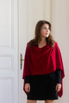 A woman wearing a deep red draped cowl-neck shawl over a black dress, standing in front of a white paneled door.