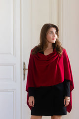 A woman wearing a deep red draped cowl-neck shawl over a black dress, standing in front of a white paneled door.