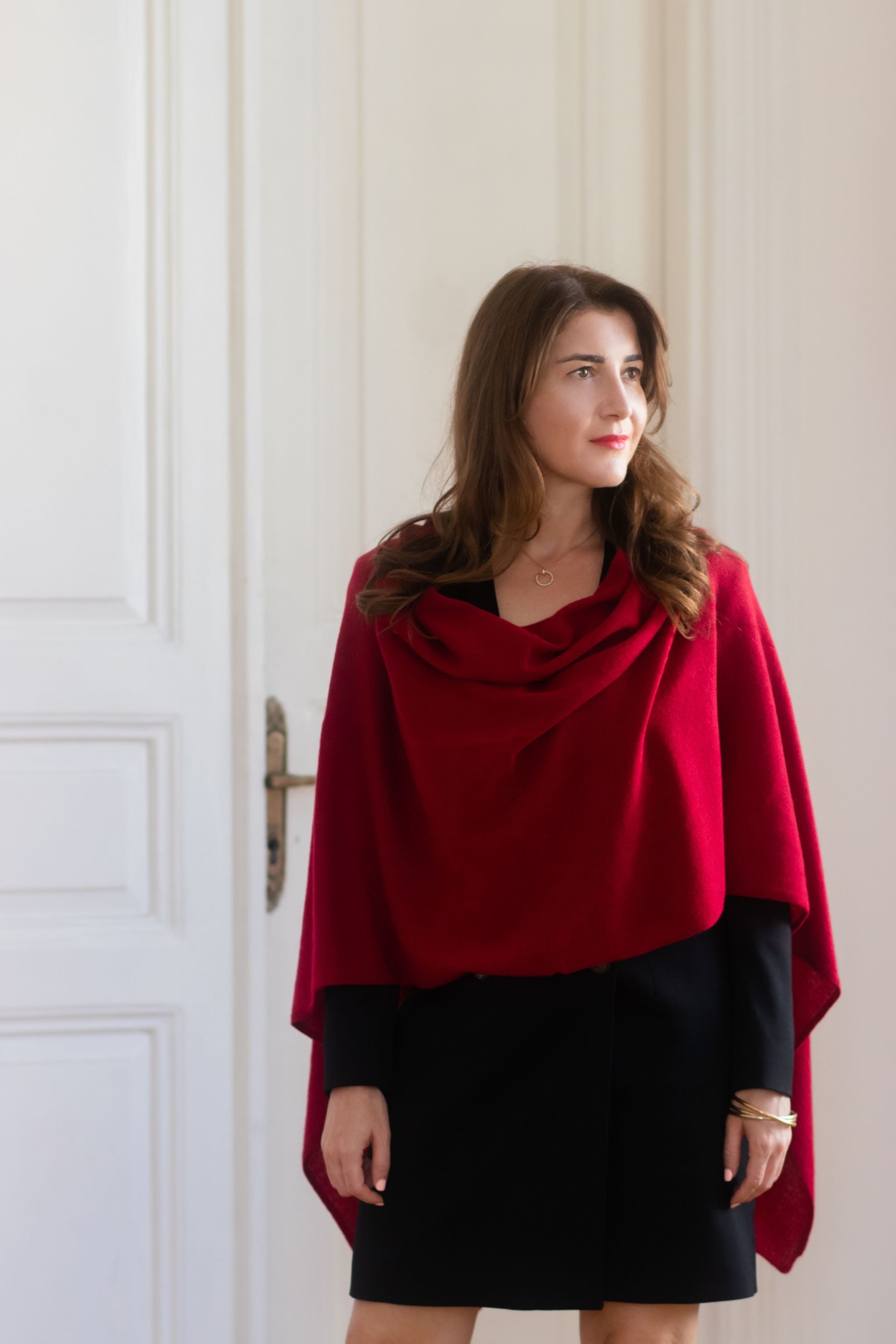 A woman wearing a deep red draped cowl-neck shawl over a black dress, standing in front of a white paneled door.
