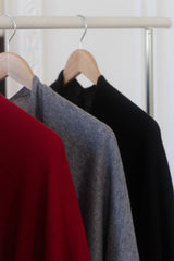 Three knit sweaters in red, gray, and black hanging on wooden hangers from a light wood clothing rack.