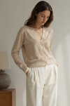 A woman wearing a light beige knit cardigan with gold buttons and white trousers, standing in a softly lit indoor setting.