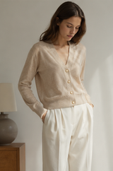 A woman wearing a light beige knit cardigan with gold buttons and white trousers, standing in a softly lit indoor setting.