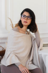 A woman with dark hair and glasses wearing a light beige knit poncho sweater over a taupe long-sleeve top, seated in a bright, elegant room.