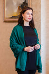 A woman wearing a dark charcoal gray knit top and a teal green cashmere wool shawl draped over her shoulders, standing in an indoor setting with a beige wall and framed picture in the background.